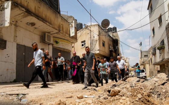 A man in a bishop's cassock walks with a large group of men between concrete buildings and rubble