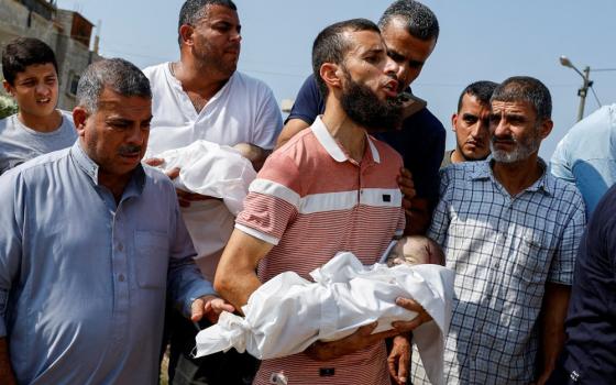 A group of brown men look sorrowful. Two carry infants wrapped in white. Blood is visible on one baby's head.