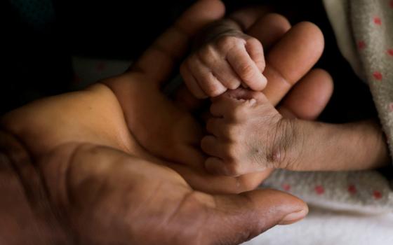 A large brown adult hand holds two incredibly skinny and small brown newborn hands and arms