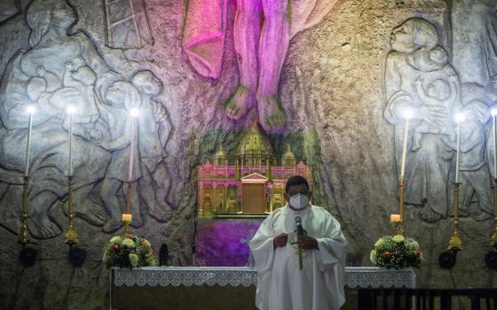 A priest wearing a face mask and white vestments holds a microphone and stands in front of a stone wall with religious carvings and purple light