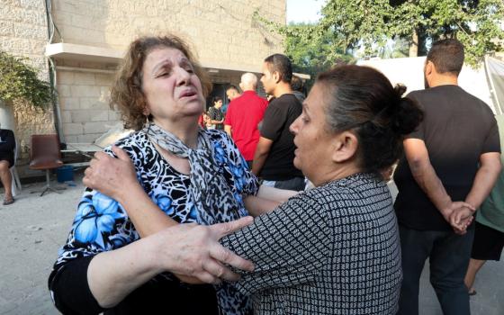 Two women, one with an extremely distressed facial expression, hold each others shoulders while standing outside among other peopel