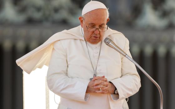 Pope Francis bows his head and folds his hands as he stands in front of a microphone and his mozzetta is blown to the side.