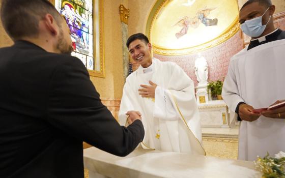 An East Asian man wearing white vestments shakes hands with another man, while another man wearing a clerical collar and a mask stands beside.