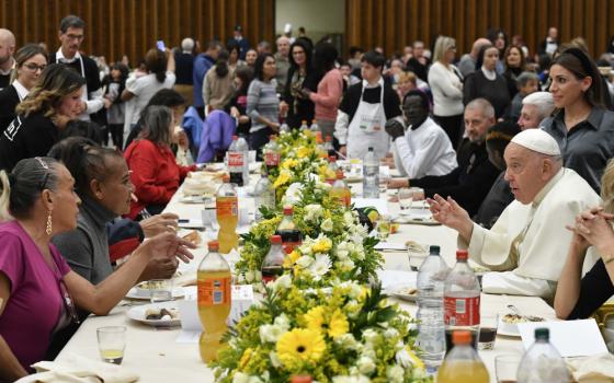 Pope Francis sits at a long table with many other people on both sides. Food, flowers and soft drinks are on the table.