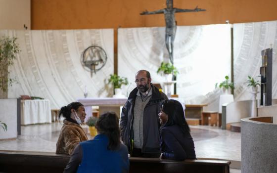 Three women sit in pews in a church while a casually dressed priest speaks with them