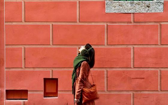 Woman wearing coat and scarf stands outside building with brick walls and looks upward. 