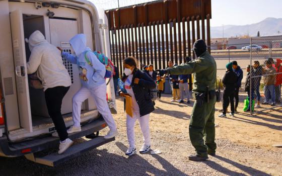 A group of people in hoodies gets the back of a van as a person wearing green with a walkie talkie points them into the van. They are beside the border wall.