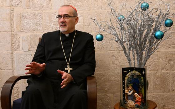 A man wearing a cassock, pectoral cross and red zucchetto sits next to a nativity scene and green orbs