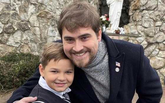 A man wearing a turtleneck and a jackets leans down to pose with a boy in a sweater vest. A statue of Mary is visible in the background.