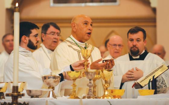 A bald man in white vestments holds a paten and speaks. Other priests in white surround him behind the altar.