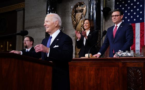 President Biden, smiling, stands at lectern, Vice President Harris and Speaker Johnson stand behind. 