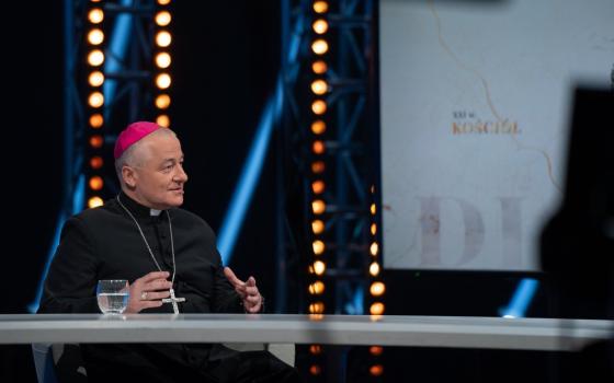 Bishop, in black cassock and purple zucchetto, talks on television. 