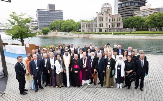 Group poses for photo before river, buildings in background. 