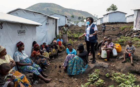 People sit on ground among tent-structures in refugee camp. 