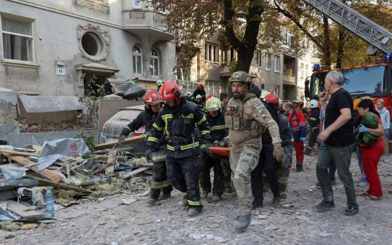 Decimated street, covered in rubble; uniformed emergency workers carry stretcher through wreckage.