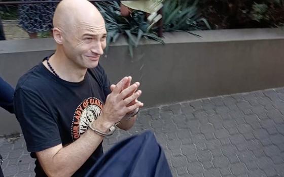 Olszewski smiles with hands folded in prayer; his head is shaven, and he is thin.