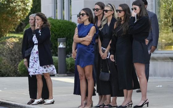 A group of women assembly, watching the hearse out of frame.