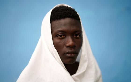 Young man, draped in white raiment, peers poignantly into camera; behind him is blue background.