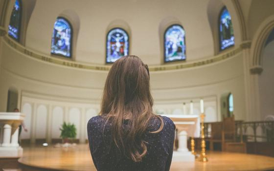 A photo shows a closeup shot of woman in the foreground, pictured inside of a church. She is facing toward the altar with her back to the camera. (Unsplash/Kenny Eliason)