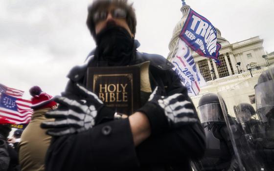 Rioter with face obscured, holds bible to chest, in the background is Capitol building dome and various flags, including one bearing former president Donald Trump's last name.