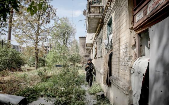 A man wearing protective gear walks along side an overgrown and crumbling building.