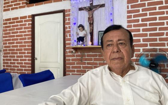 Rabanales sits at a table, behind him is a crucifix and the El Nino Dios dressed as a migrant in a small wall shrine.