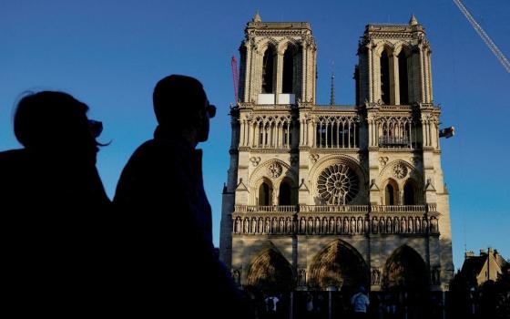 The Cathedral looms against blue sky; on right side is the arm of a crane; in left third of frame are two silhouetted figures of bystanders.