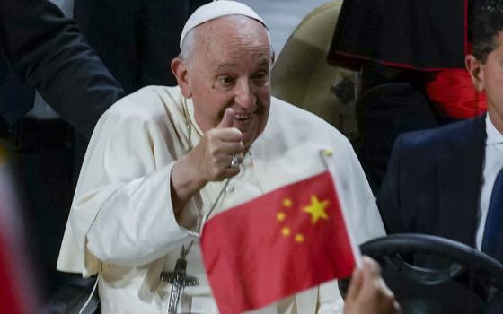 Francis smiles and gives thumbs up, foregrounded is small, hand-held Chinese flag.