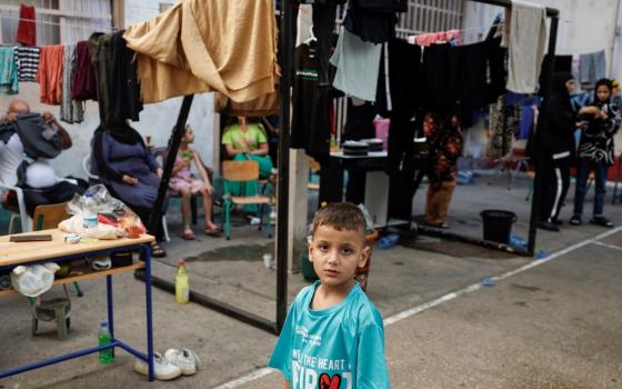 Young boy wearing oversized shirt foregrounded; in background are other people, and a metal frame draped with cloth.