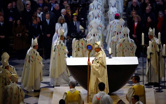 Archbishop, fully vested, pictured in profile holding crozier with massive blue stone; other bishops process, and seated in front of right row are France's presidential couple.