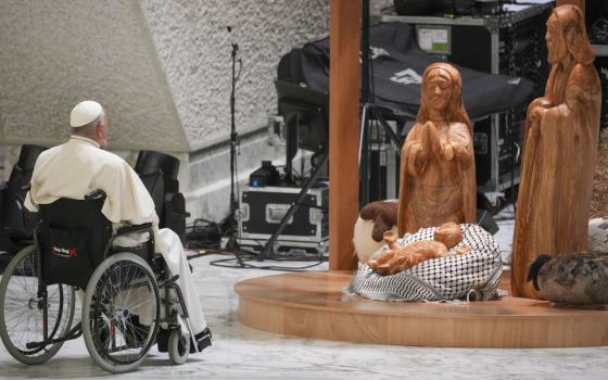 Pope Francis in wheelchair sits before wooden nativity scene with keffiyeh.