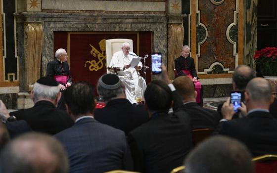 Pope sits on raised dais, people seen watching.