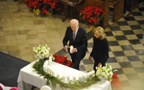 President Biden and First Lady Jill Biden wear black and approach altar adorned with greenery and candles. The president holds large white pillar candle. 