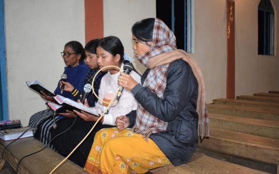 Dkhar, sits on bench in church singing into microphone.