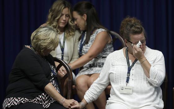 Four women sit while dabbing tears and holding hands.