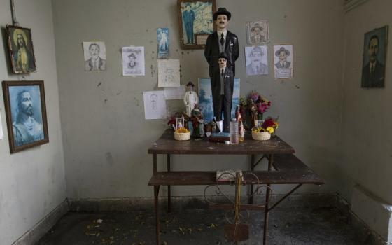 A large colored statue of Hernandez stands ont ables with flowers and other religious articles in a small gray room; walls covered with pictures and drawings of both Jesus Christ an Hernandez. 