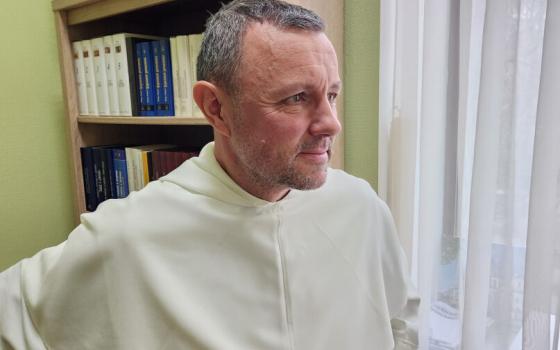 The priest stands wearing white habit staring out of window. 