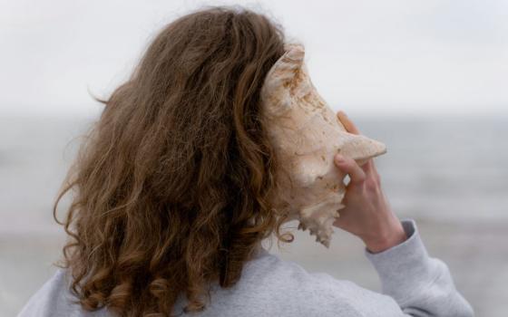 Woman faces ocean and holds shell to her ear.