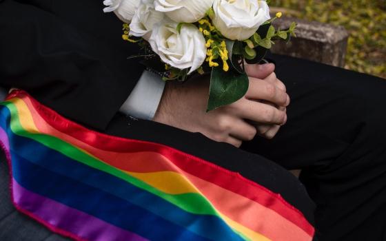 Rainbow flag next to seated man in black suit holding a bouquet of white roses