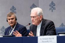 Steiner shown sitting and blue-clothed press conference table speaking into microphone; behind is a background emblazoned with the Vatican's coat-of-arms.