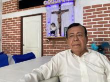 Rabanales sits at a table, behind him is a crucifix and the El Nino Dios dressed as a migrant in a small wall shrine.