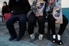 Three people with faces out of frame sit on a bench, two hold paper bags.